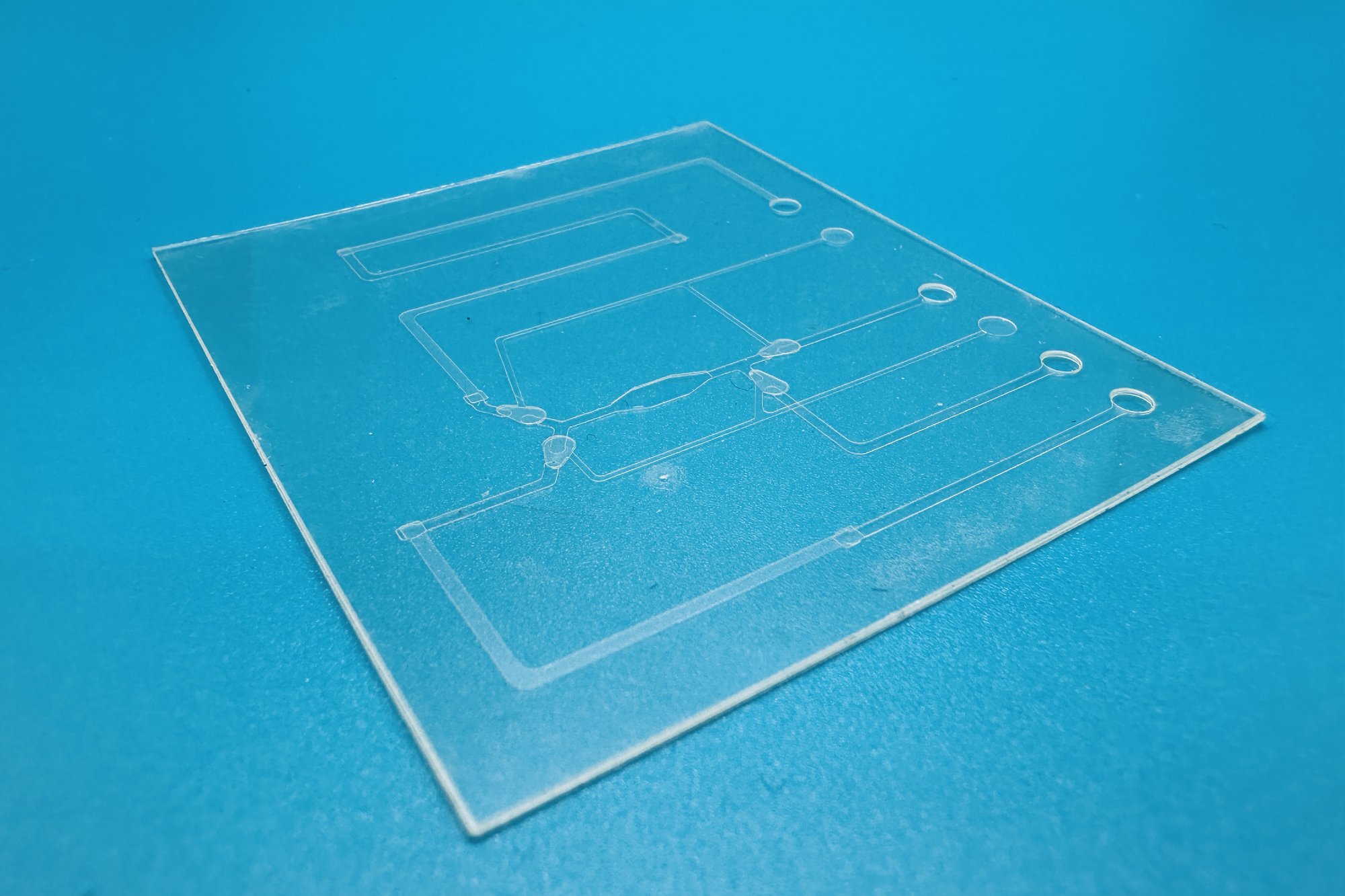 Clear microfluidic metering chip with laser-made channels and ports, on a blue background