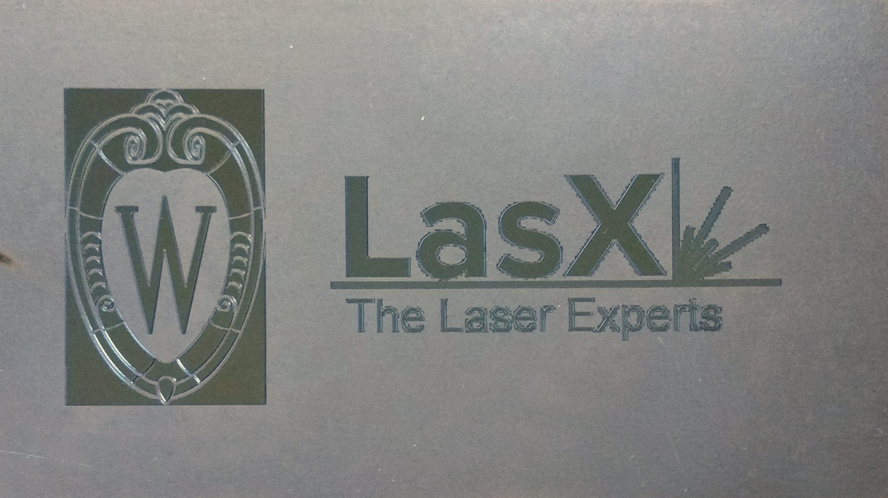 Steel coupon with the LasX wordmark and a University of Wisconsin Motion W laser-polished to a mirror finish against the surrounding matte ground surface